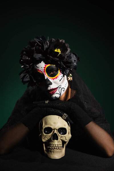 Woman in mexican halloween costume touching skull on dark green background 