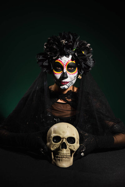 Woman in wreath with veil and catrina makeup looking at camera near skull on dark green background 