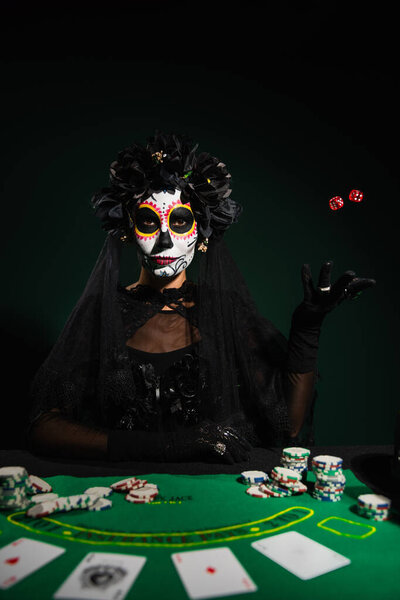 KYIV, UKRAINE - SEPTEMBER 12, 2022: Woman in santa muerte makeup and costume throwing dice near playing cards on dark green background 
