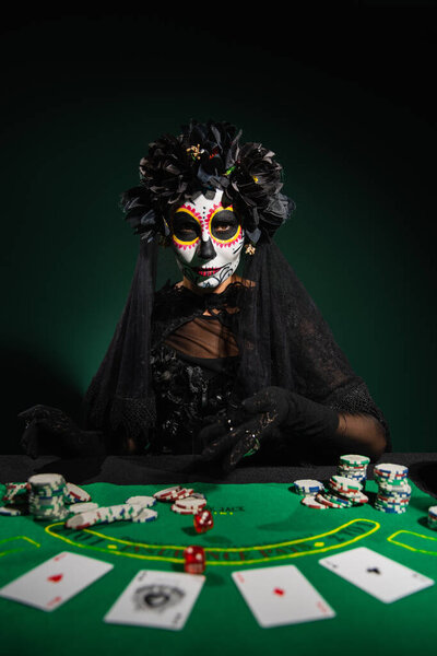 KYIV, UKRAINE - SEPTEMBER 12, 2022: Woman in mexican day of dead costume throwing dice on table on dark green