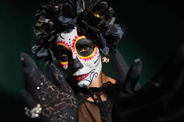 Woman in sugar skull makeup and wrench with black flowers looking at camera on dark green background