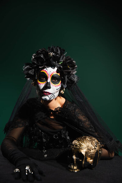 Woman in santa muerte costume and makeup looking at camera near skull isolated on dark green 