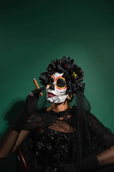Woman in mexican day of death creepy costume holding cigar on green background