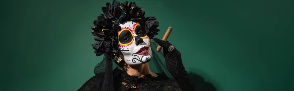 Woman with sugar skull makeup and black wreath holding cigar on green background, banner