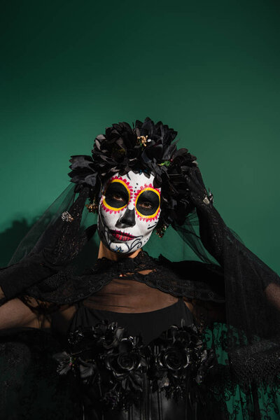 Woman in halloween santa muerte costume and wreath looking at camera on green background 
