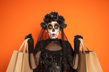 Geleneksel ölü kostümü ve turuncu üzerine izole edilmiş alışveriş torbalarını gösteren kedi makyajlı kadın.