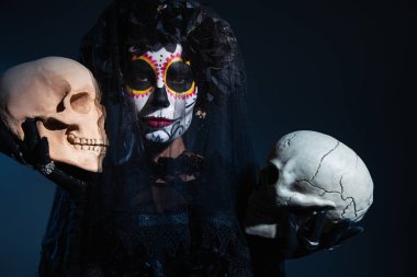 Siyah cadılar bayramı kostümü giymiş, elinde kafatasları olan ve koyu mavi arka planda kameraya bakan bir kadın.