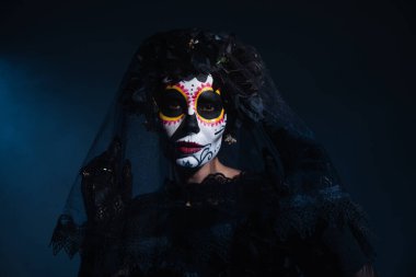 woman in mexican day of dead costume and catrina makeup looking at camera under black lace veil on dark blue background clipart