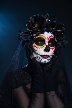 Siyah çelenkli kadın, koyu mavi arka planda geleneksel Santa Muerte makyajıyla el ele tutuşuyor.