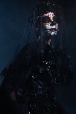 woman in traditional santa muerte makeup and black lace veil looking at camera on dark blue background clipart