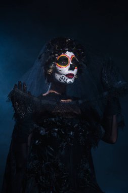 woman in witch costume and sugar skull makeup holding lace veil on dark blue background clipart