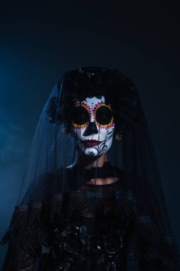 woman in creepy halloween makeup and black wreath with veil posing with closed eyes on dark blue background clipart