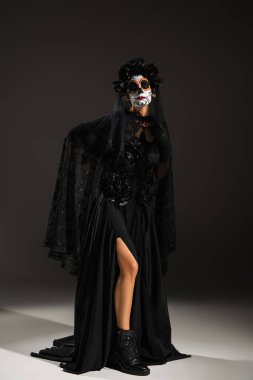 full length of woman in sugar skull makeup posing in black halloween costume on dark background clipart
