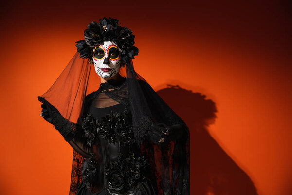 Woman in halloween catrina makeup touching black veil on red background 