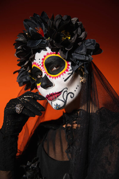 woman in black wreath and spooky halloween makeup holding hand near face and looking at camera on orange background
