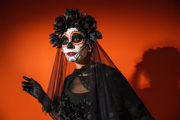 woman in mexican day of dead makeup and black spooky costume looking away on orange background with dark shadow