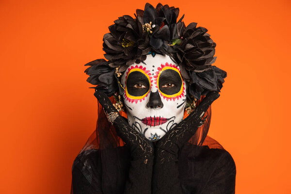 portrait of woman in black wreath and sugar skull halloween makeup posing with hands near face isolated on orange
