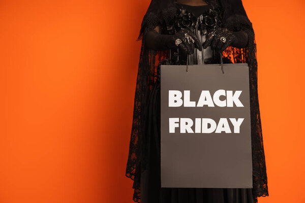 cropped view of woman in spooky halloween makeup holding shopping bag with black friday lettering isolated on orange