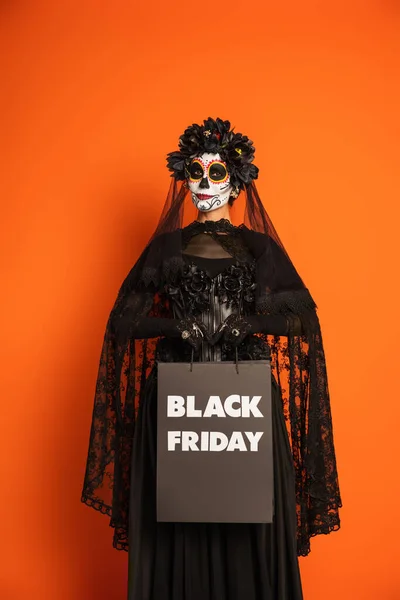 woman in catrina makeup and black costume with veil holding black friday shopping bag isolated on orange