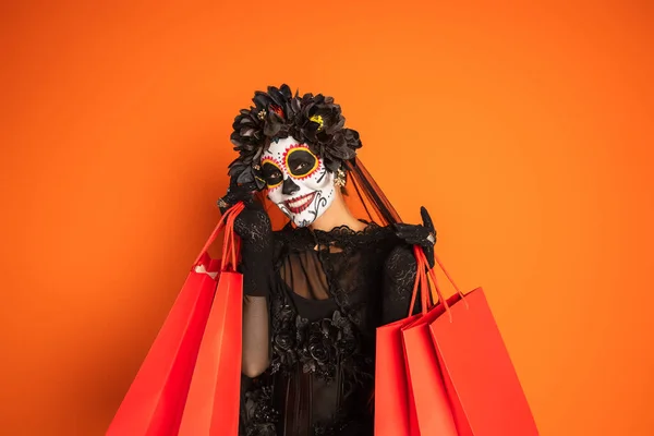 ブラック魔女の衣装を着た楽しい女性とオレンジに孤立したショッピングバッグを示す不気味なハロウィンメイク — ストック写真