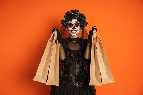 ブラック魔女の衣装を着た陽気な女性とオレンジに孤立したショッピングバッグを示すハロウィンメイク — ストック写真