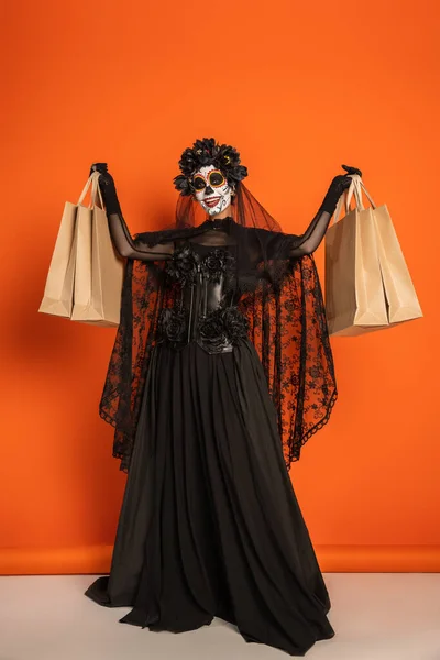 黒い魔女のドレスとハロウィンの化粧を着た陽気な女性の完全な長さオレンジの背景にショッピングバッグを示す — ストック写真
