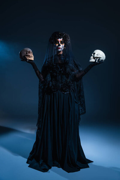 full length of woman in black costume and spooky halloween makeup holding skulls on dark background with blue light