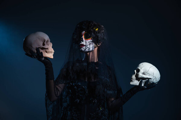 woman in traditional mexican day of dead makeup and costume holding skulls on dark blue background