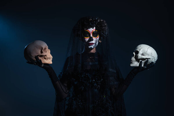front view of woman in halloween makeup and black costume standing with skulls on dark blue background