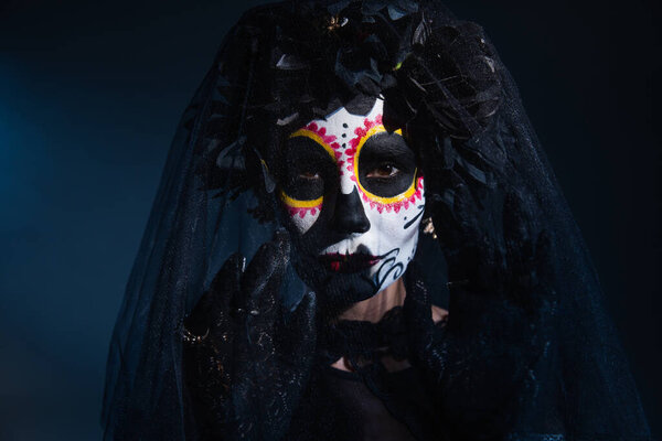 woman in sugar skull halloween makeup gesturing near black veil on dark blue background