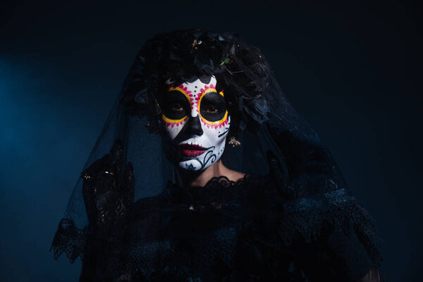 woman in mexican day of dead costume and catrina makeup looking at camera under black lace veil on dark blue background