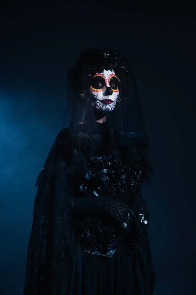 woman in scary halloween makeup and black costume with veil on dark blue background