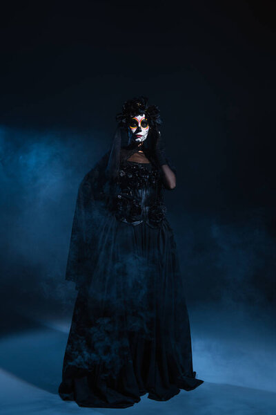 full length of woman in black costume and creepy halloween makeup posing with hand on waist on dark background with blue fog