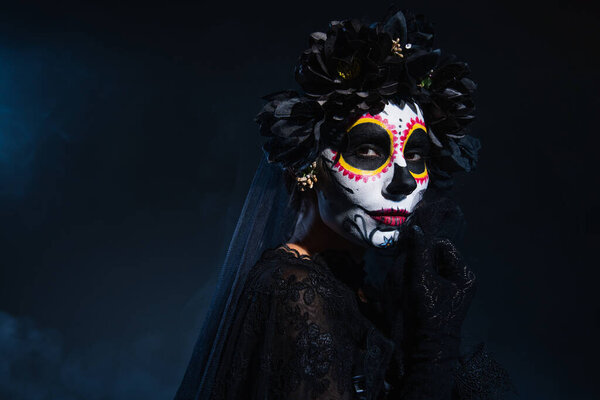 woman in traditional santa muerte makeup and black wreath holding hand near face on dark blue background