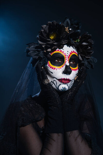 woman in black wreath holding hands near face with traditional santa muerte makeup on dark blue background