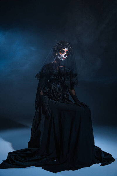 woman in halloween makeup and black witch dress sitting on dark smoky background with blue light