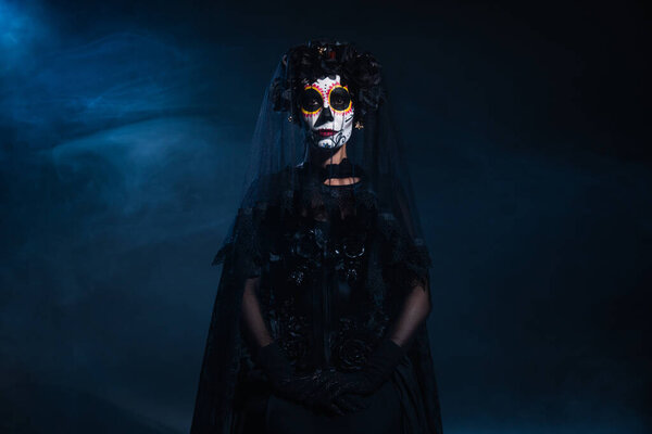 woman in catrina makeup and black costume with veil on dark background with blue fog