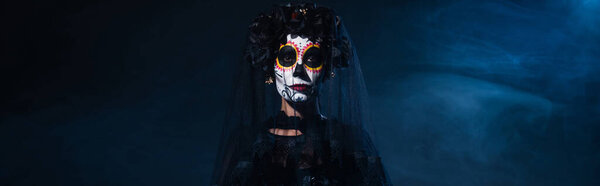 woman in mexican day of dead makeup and black wreath with veil on dark background with blue smoke, banner