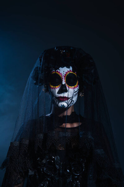 woman in creepy halloween makeup and black wreath with veil posing with closed eyes on dark blue background