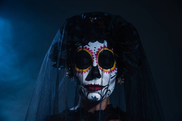portrait of woman with closed eyes and scary halloween makeup wearing black wreath and veil on blue foggy background