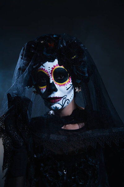 portrait of woman in mexican day of dead makeup and black costume looking at camera on dark blue background