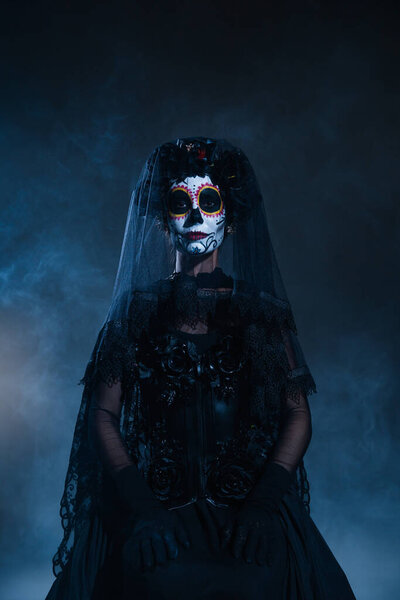 front view of woman in scary halloween makeup and black costume with veil on dark blue foggy background