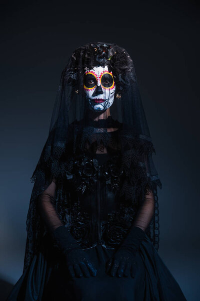 front view of woman in black costume and traditional santa muerte makeup sitting on dark blue background