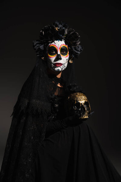 woman in scary halloween makeup and dark costume holding golden skull on black background