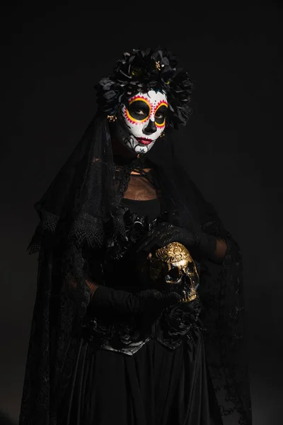 woman in creepy costume and catrina makeup holding golden skull isolated on black