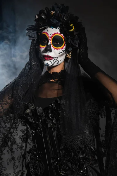 woman in sugar skull makeup and costume with black wreath and lace veil on dark background with smoke