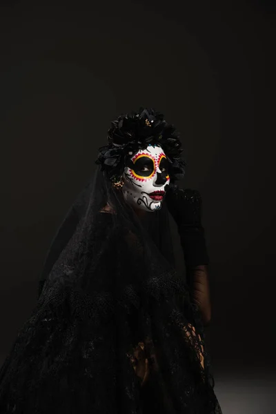 woman in spooky halloween makeup and black wreath with veil looking at camera on dark background