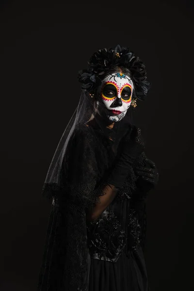 woman in sugar skull makeup and dark halloween costume with veil looking away isolated on black