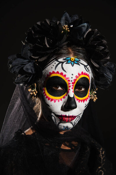 portrait of woman in halloween sugar skull makeup and dark wreath with veil looking at camera isolated on black