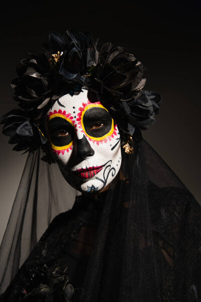 portrait of woman in halloween sugar skull makeup and black wreath with veil on dark background